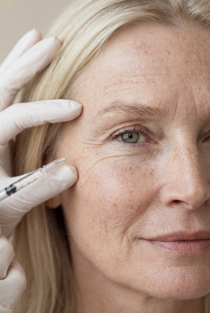 A woman with long, blonde hair and visible freckles is having a syringe held near her eye by a person wearing white gloves, indicating an aesthetic procedure.