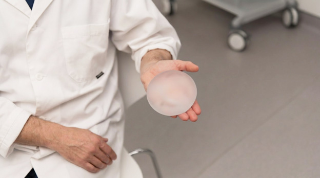 A person in a white coat holding a translucent round silicone breast implant in their palm.