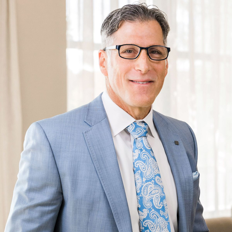 A man wearing glasses and a light blue suit, with a white shirt and a blue paisley tie, smiling at the camera.