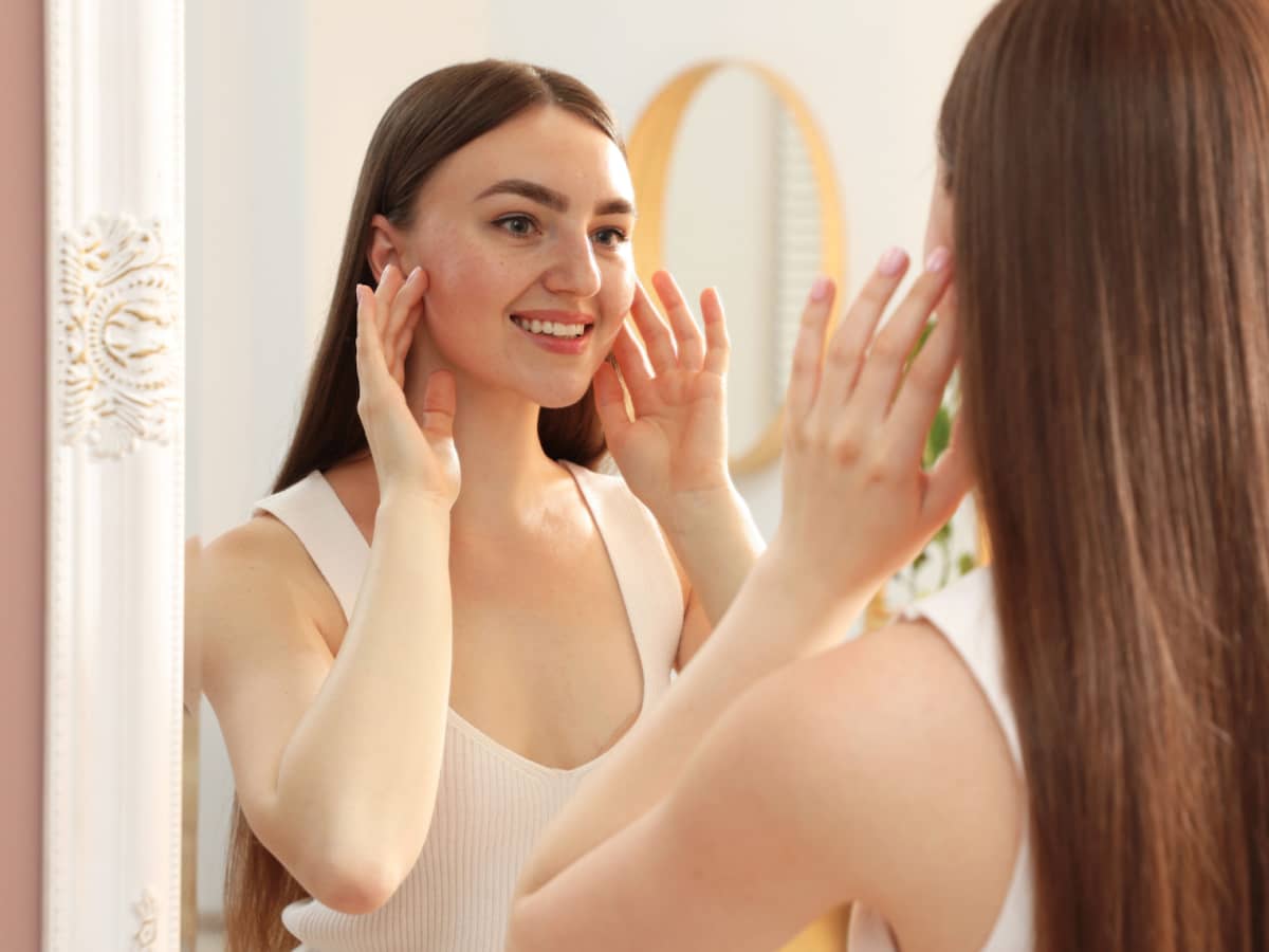 Woman smiling at her reflection in a mirror with both hands on her face