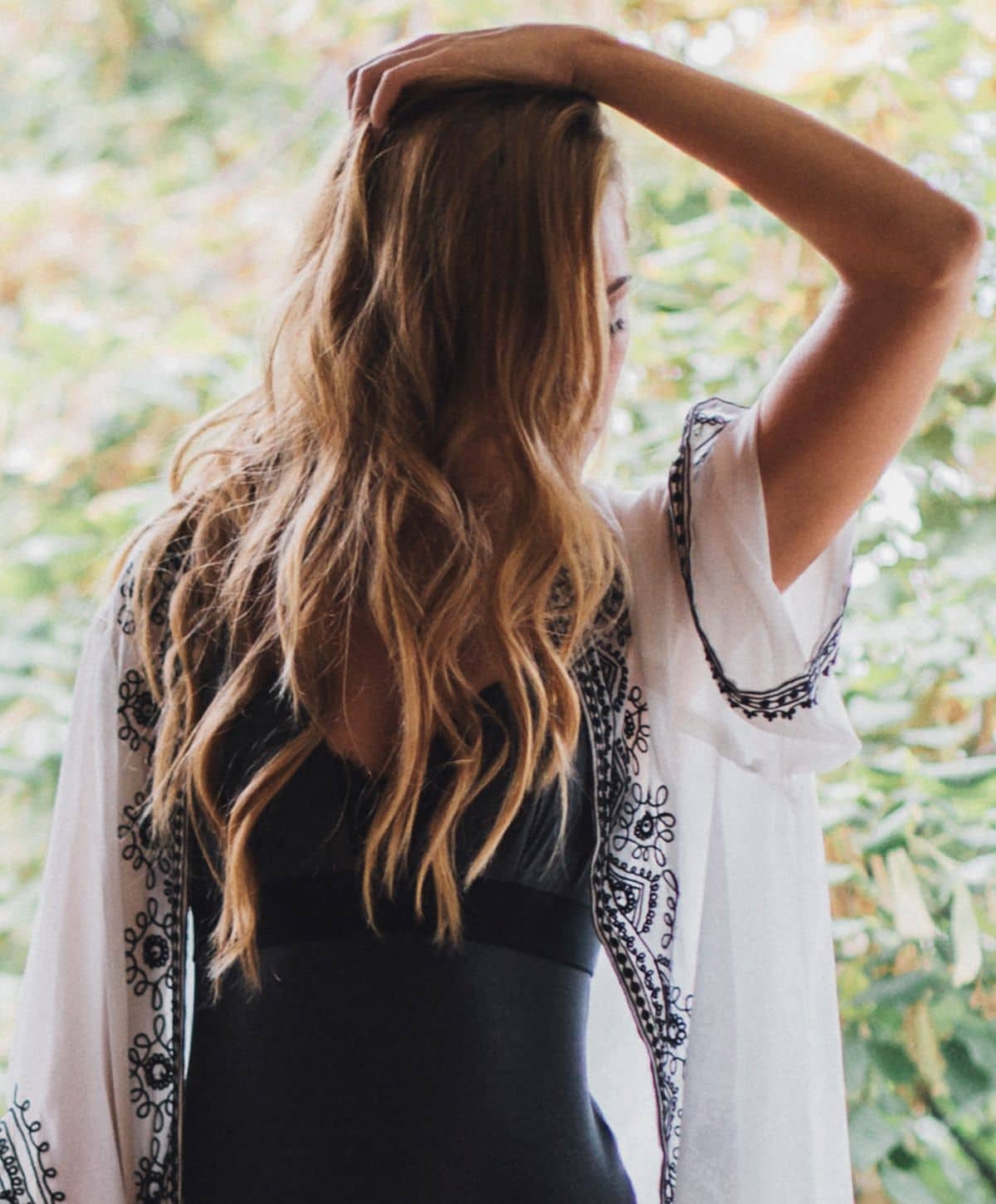 A woman with long, wavy hair wearing a black swimsuit and a white cover-up with decorative black patterns, holding her hand on her head while looking away.