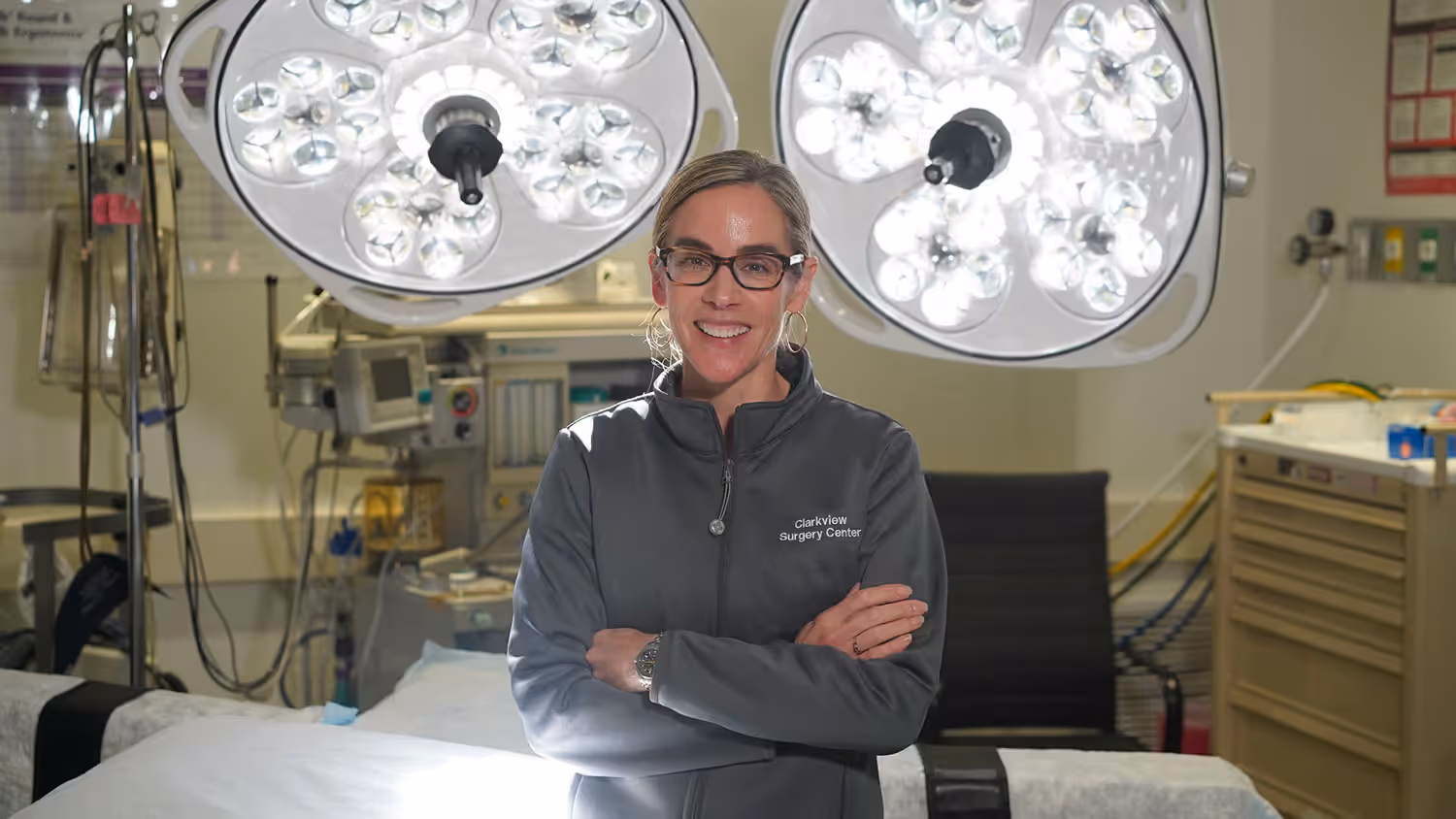 A woman with glasses smiles while standing in a medical environment, wearing a dark jacket with 'Caroline Surgery Center' text on the left chest, with overhead surgical lights in the background.
