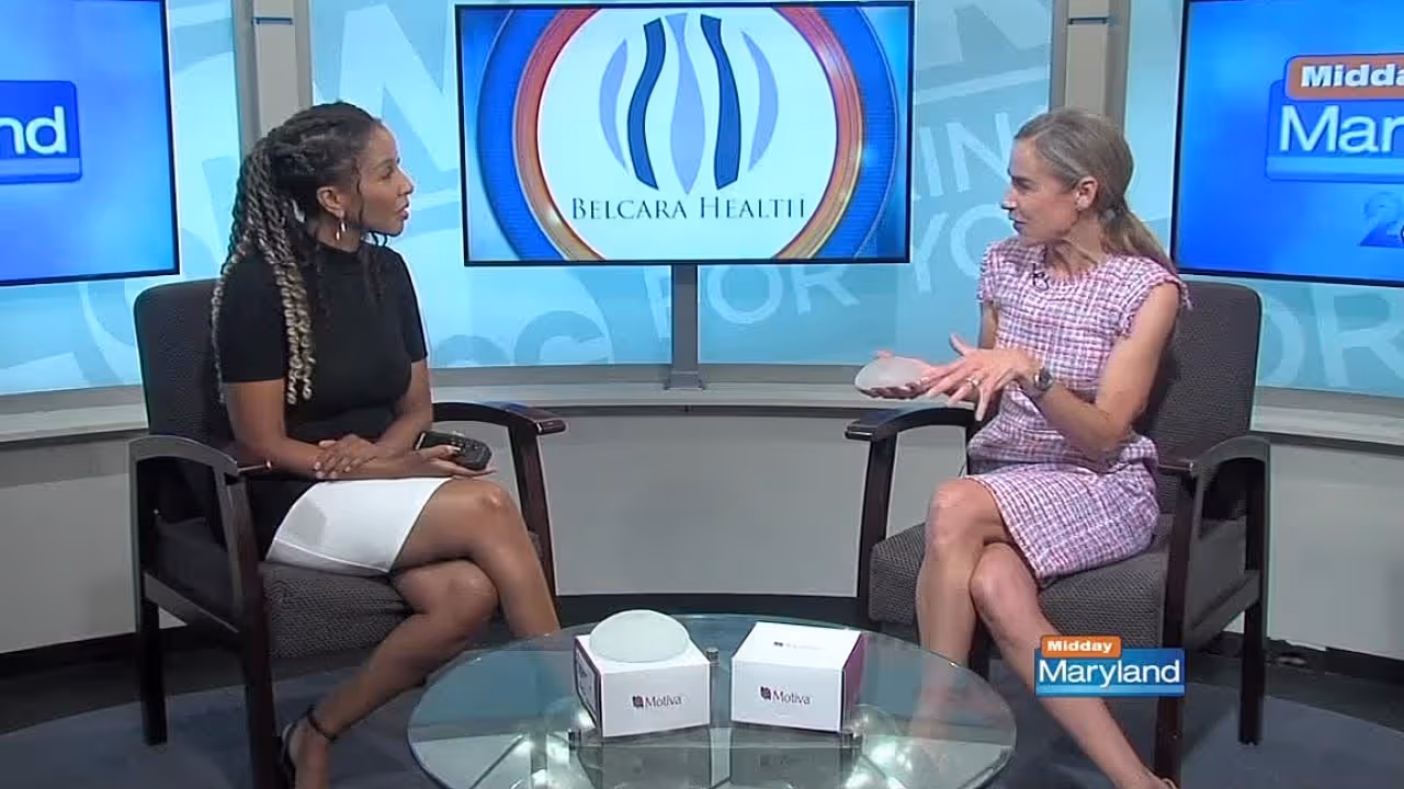 Two women engaged in a discussion, with one woman holding a breast implant and the other gesturing. In the background, a logo for Belcara Health is prominently displayed.
