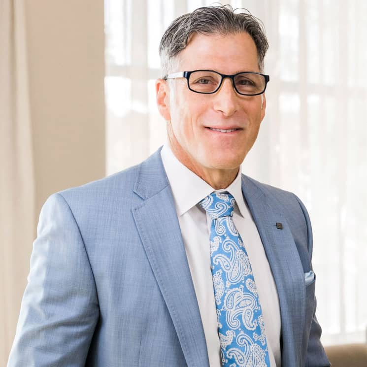 A man wearing glasses and a light blue suit, with a white shirt and a blue paisley tie, smiling at the camera.