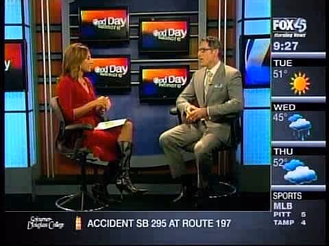 A woman in a red dress and black knee-high boots sitting on a stool while interviewing a man in a gray suit and glasses, who is also seated on a stool. They are discussing a topic related to cosmetic surgery, with a weather and news ticker visible on the.