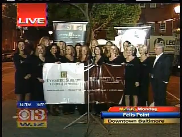 A group of women standing together in formal attire, holding a large banner with the text 'Cosmetic Surgery Center of Maryland' and 'Live' at the top left. Several promotional signs are visible in the background.