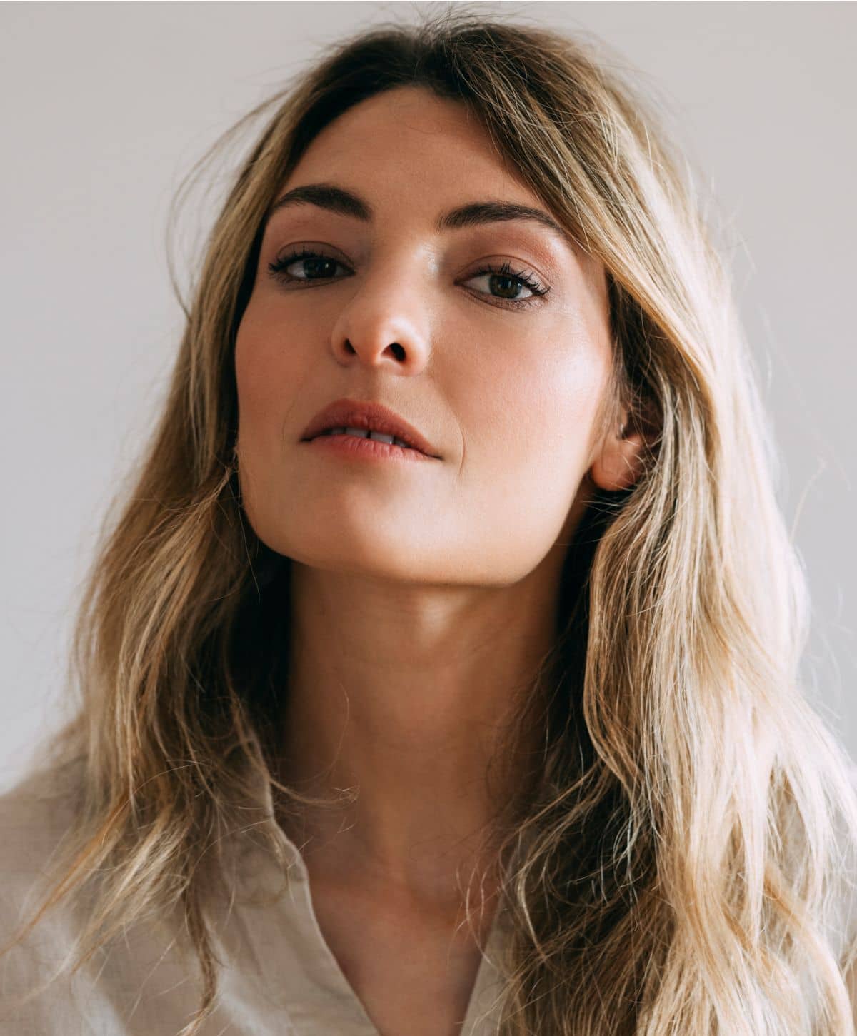 A portrait of a woman with long, wavy hair, looking slightly upwards with a neutral expression. She has a natural makeup look, and is wearing a light-colored blouse.