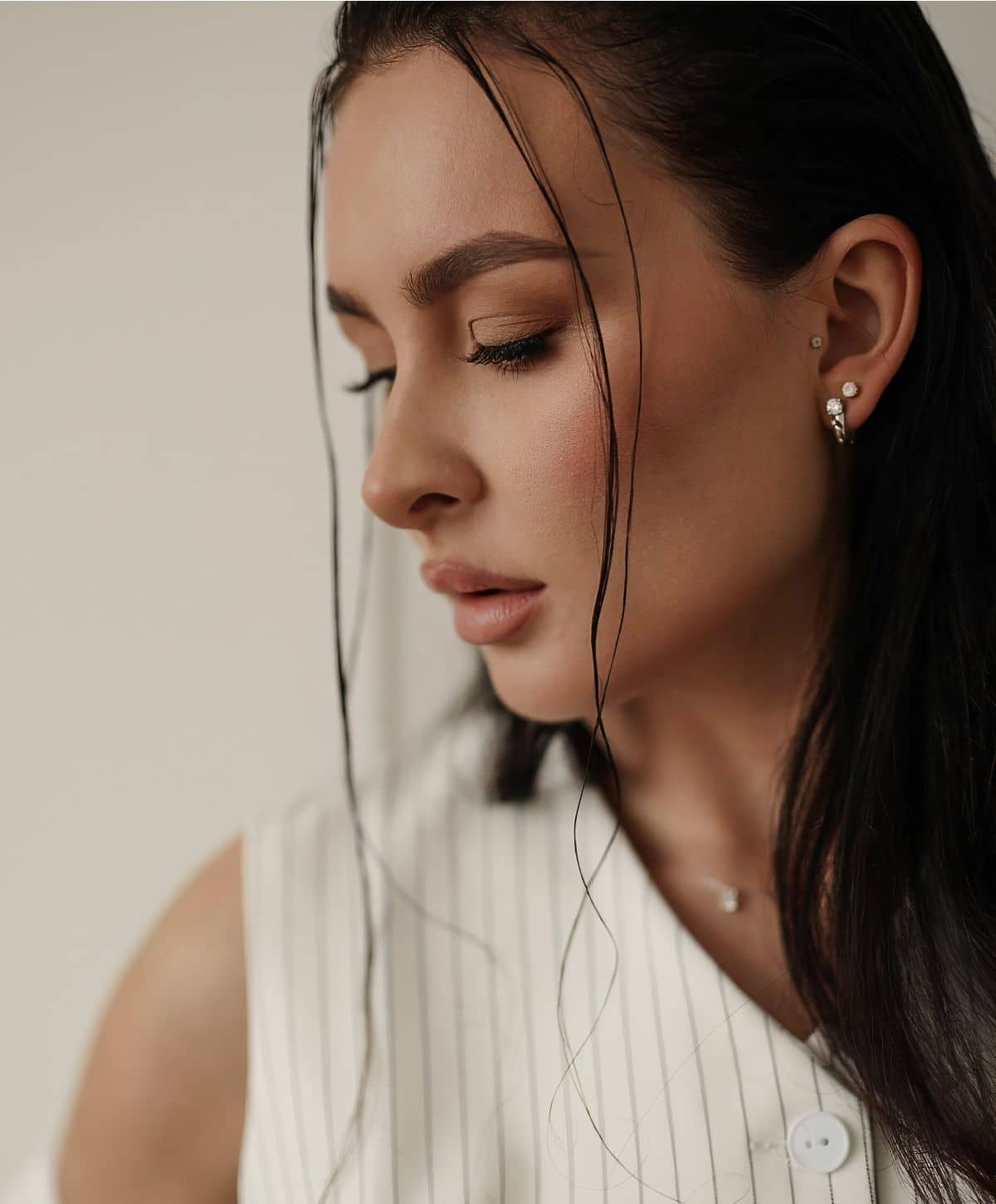 A profile portrait of a woman with long dark hair partially covering her face, wearing a striped white blouse. She has a subtle makeup look and is adorned with a small earring.