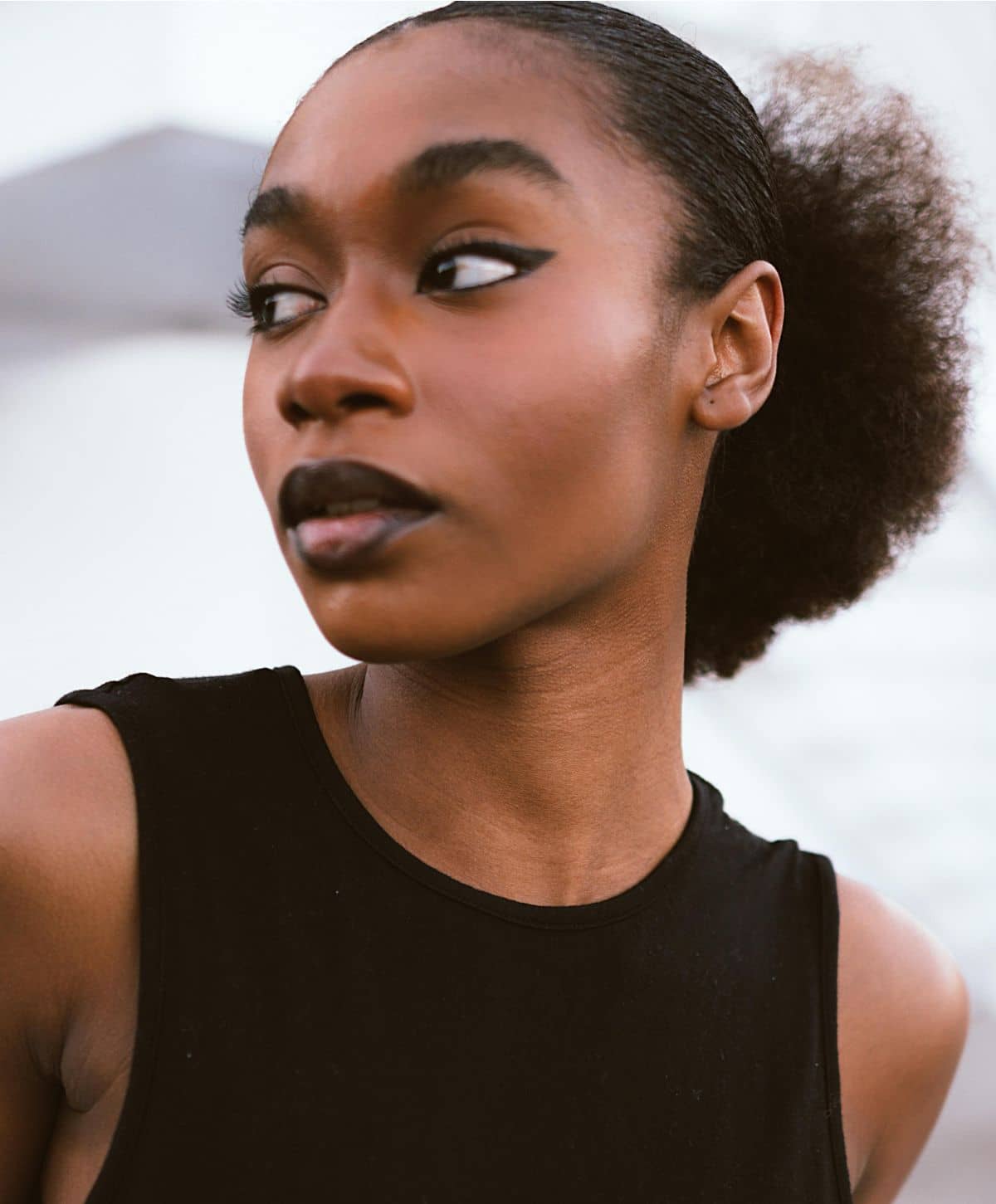A woman with a sleek hairstyle wearing black lipstick and a sleeveless dark top, gazing over her shoulder with an expressive face.