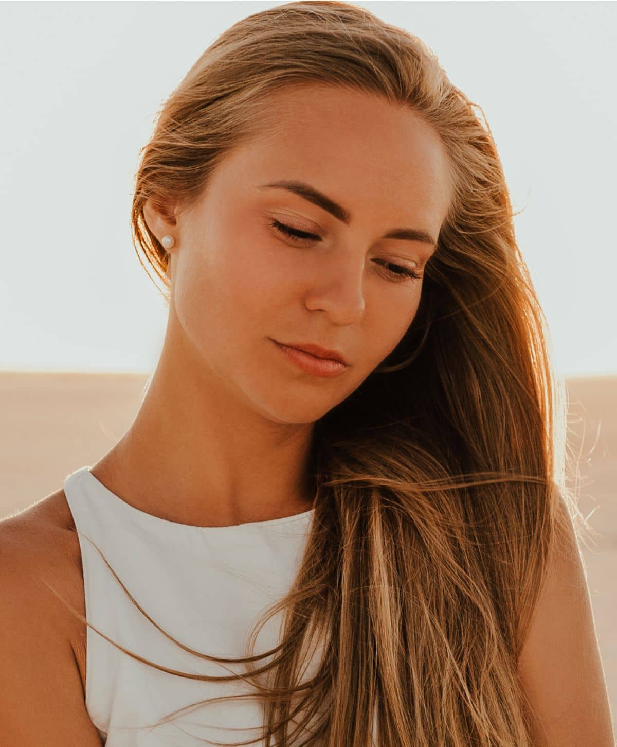 A woman with long, flowing hair, wearing a white top, looking down with a soft expression.