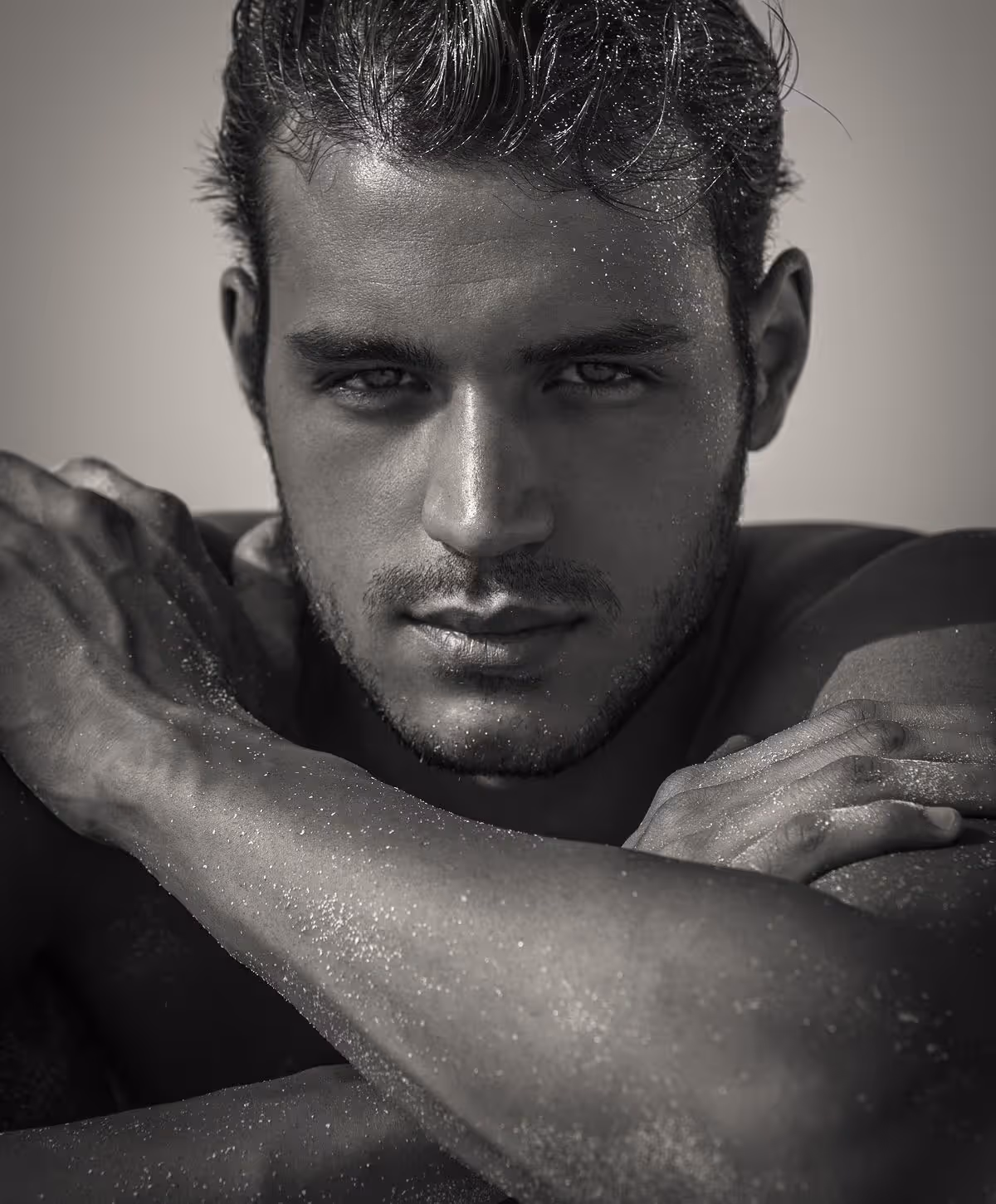 A close-up of a male model with tousled hair, showcasing a serious expression and muscular arms crossed in front of his chest, glistening with water droplets, set against a soft focus background.