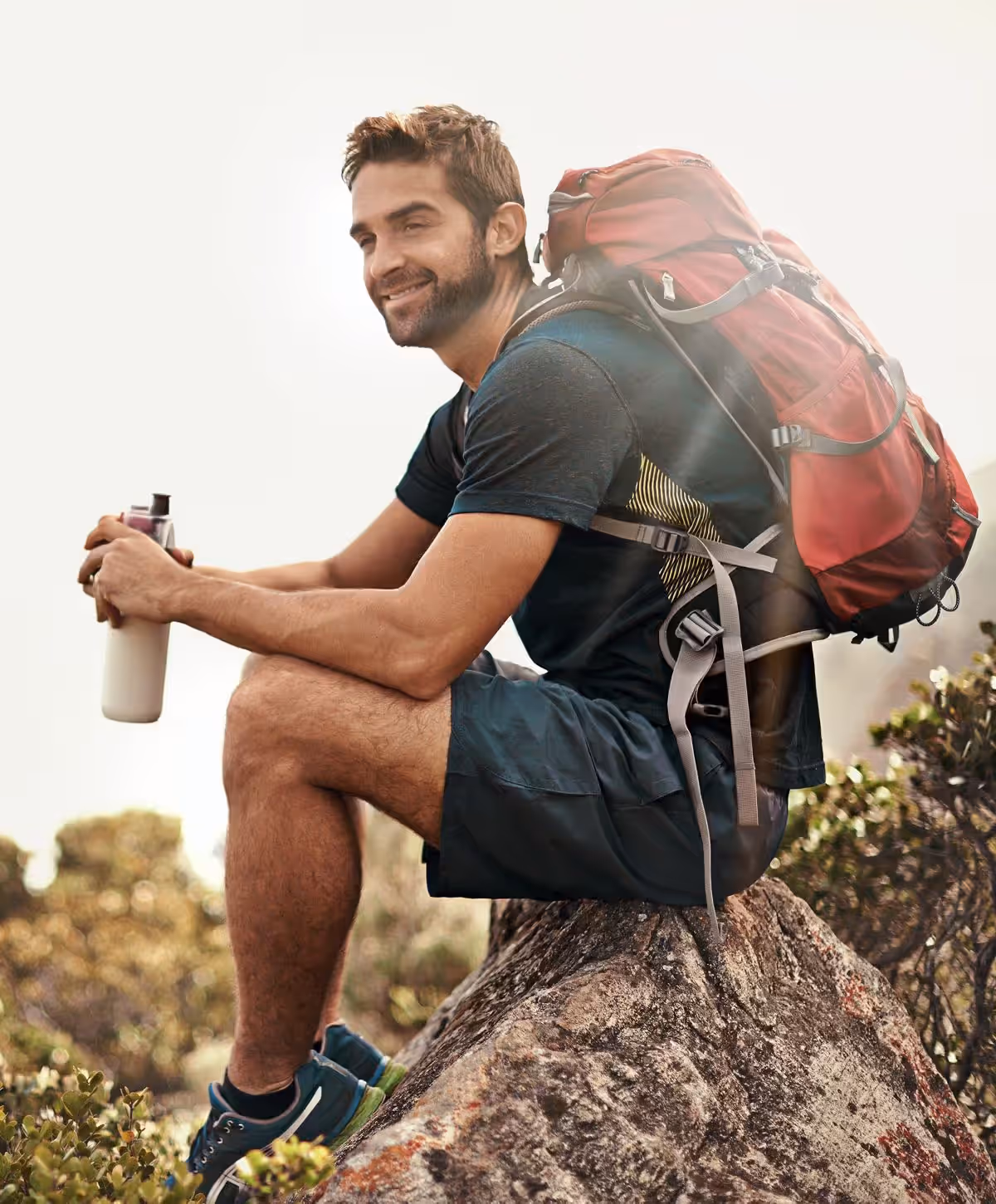 A man with a backpack sitting on a rock, holding a water bottle, smiling while wearing a short-sleeved athletic shirt and shorts.