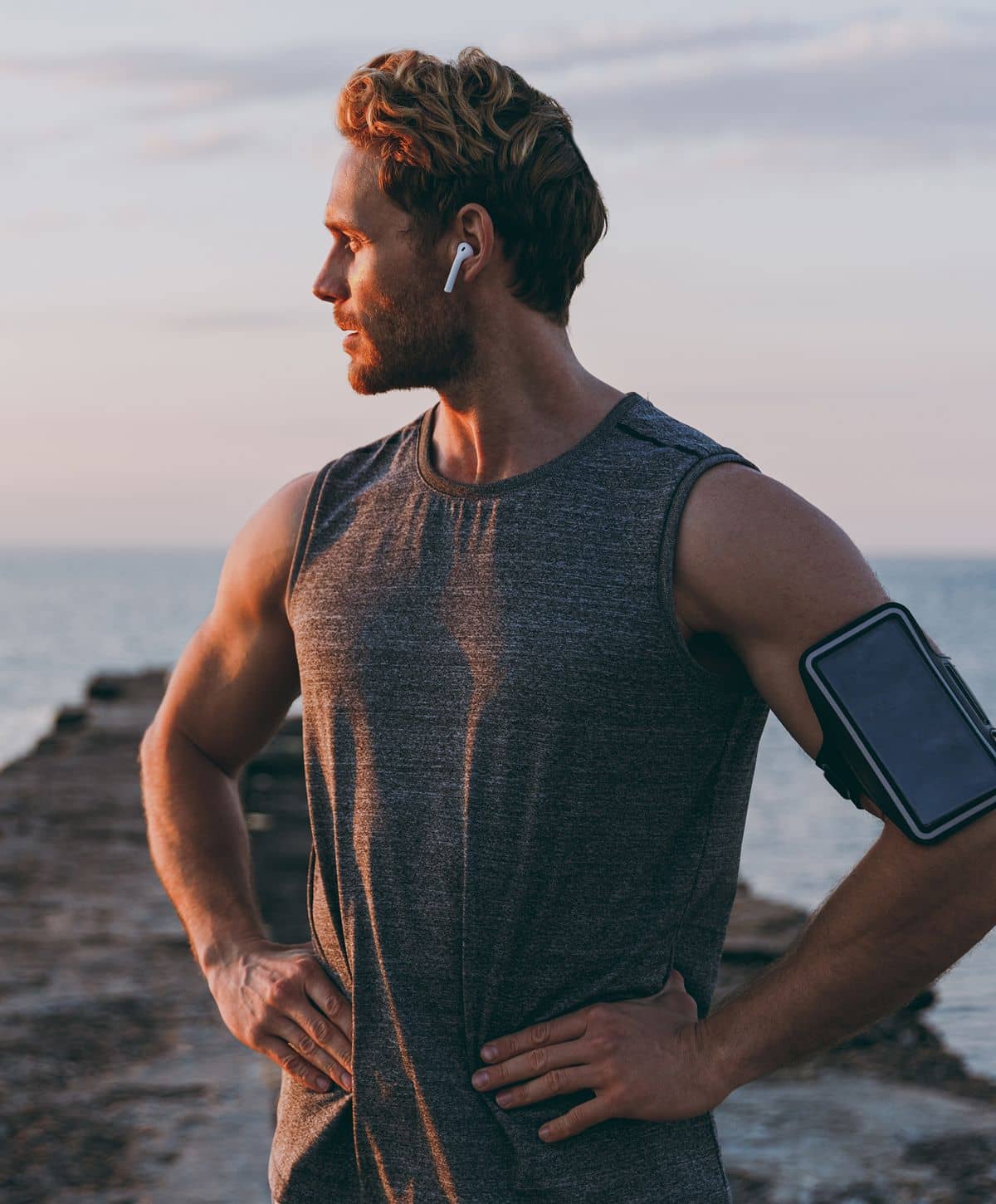 A man in a sleeveless gray athletic shirt stands on a rocky pier, looking off to the side. He wears wireless earbuds and has a phone armband on his left arm.