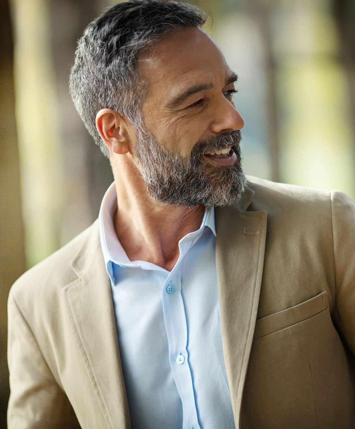 A smiling man with a beard wearing a light blue dress shirt and a tan blazer.