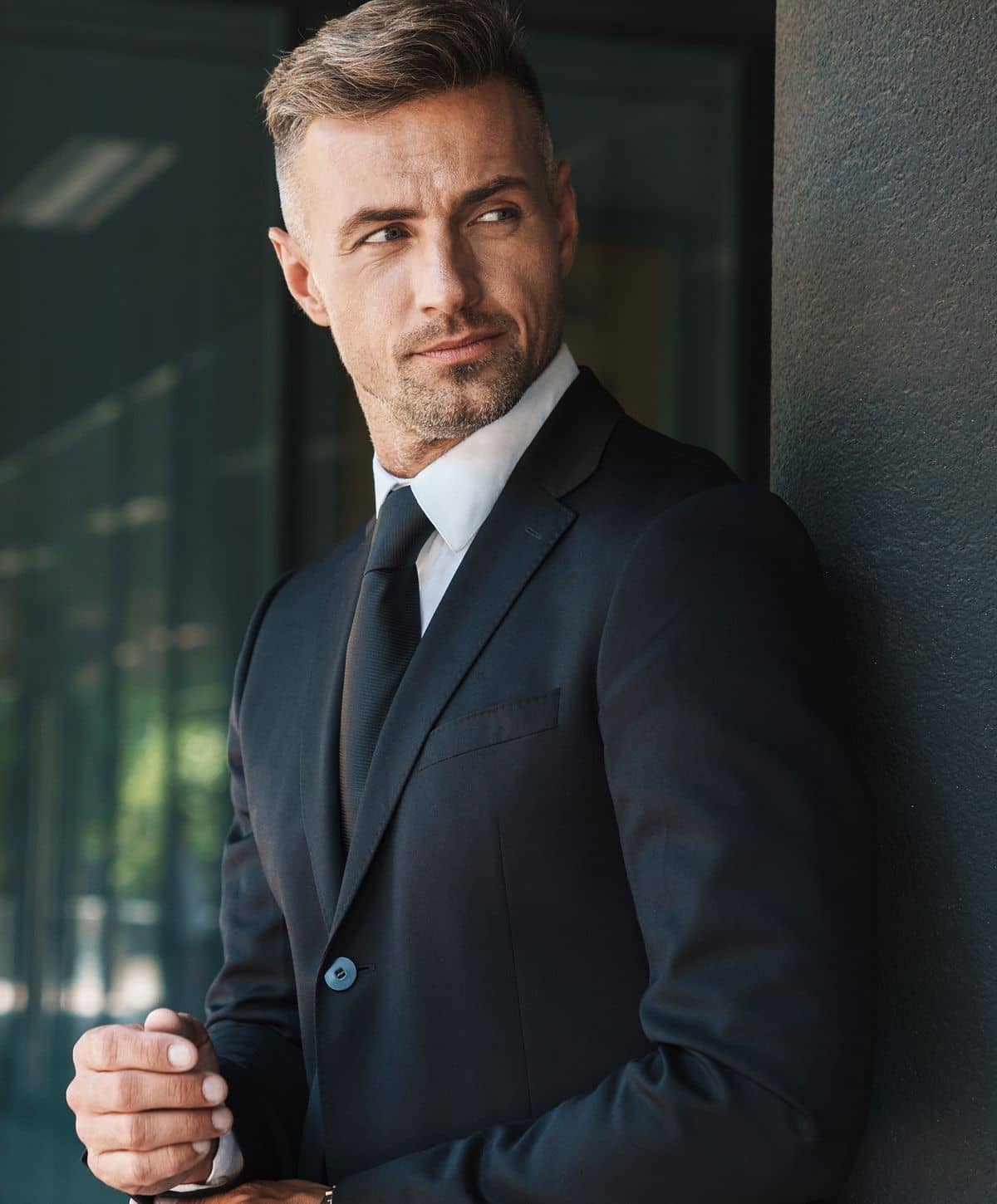 A man in a black suit and tie, standing with his arms crossed, looking off to the side with a subtle smile.