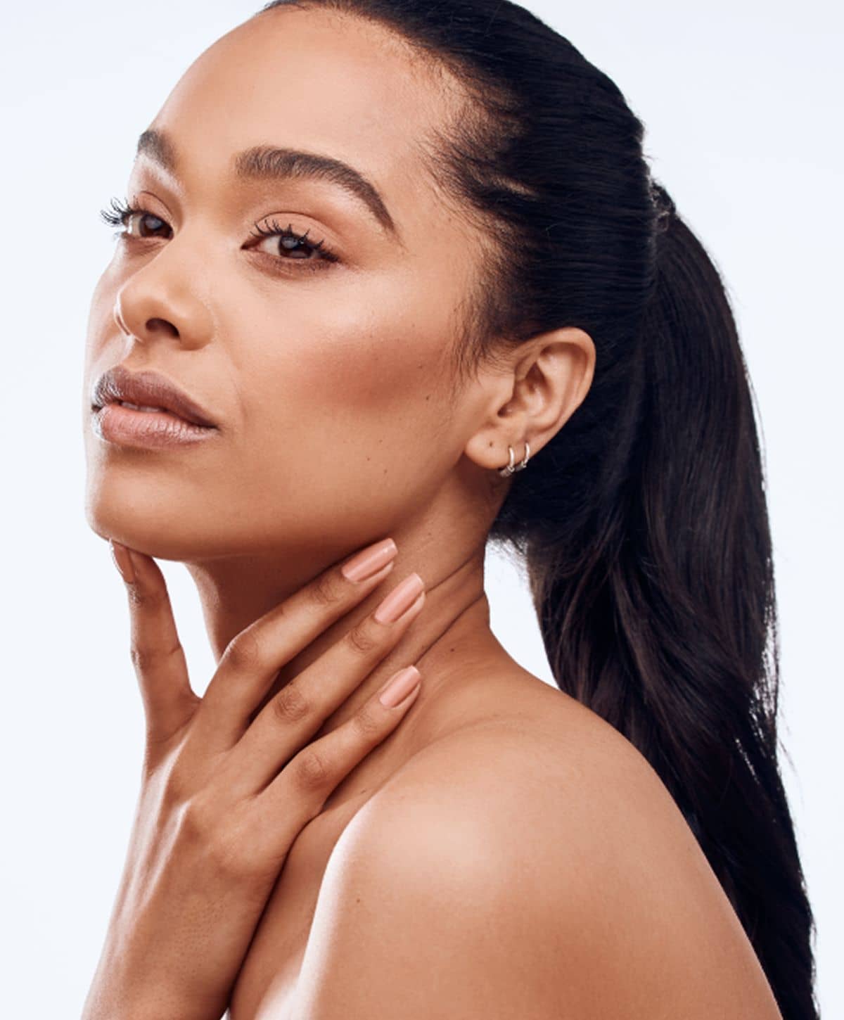 A woman with long dark hair pulled back into a ponytail, gently touching her neck with one hand while looking to the side. She has a natural makeup look and a relaxed expression.
