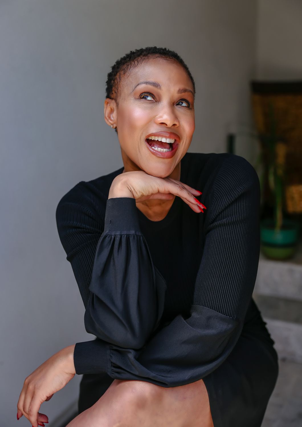 A woman with short, curly hair is smiling while resting her chin on her hand, wearing a black long-sleeve dress.