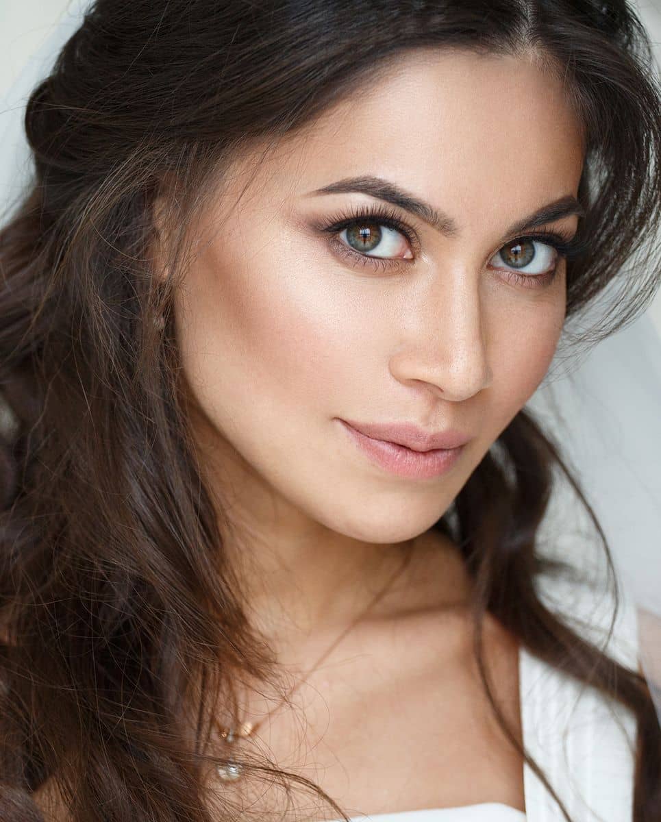 A close-up portrait of a woman with long, wavy dark hair and light brown skin. She has striking green eyes, subtle makeup, and a gentle smile.