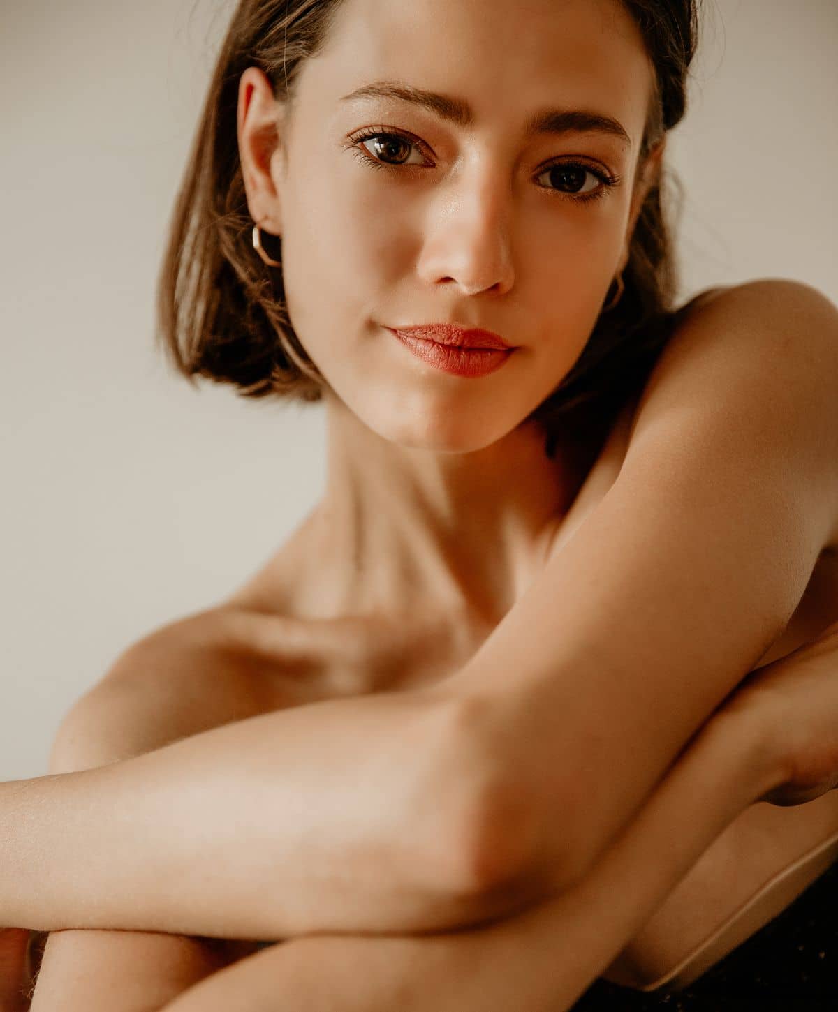 A young woman with short brown hair, wearing minimal makeup and a black top, gazes softly at the camera with her arms crossed over her body.