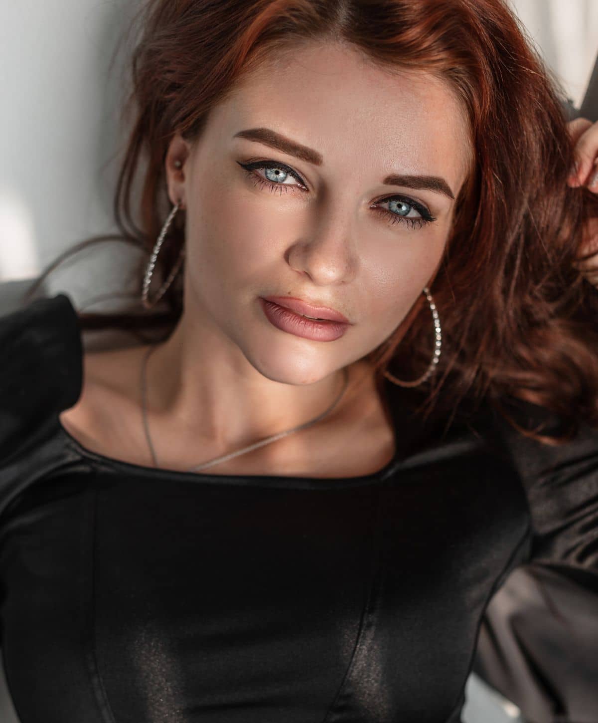 A woman with long, wavy red hair and blue eyes, wearing a black top, poses for the camera with a subtle smile and silver hoop earrings.