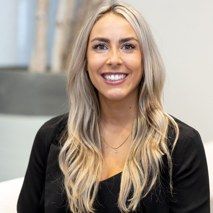 A portrait of a smiling woman with long, blonde hair, wearing a black top. She is sitting in a well-lit environment, highlighting her natural features.