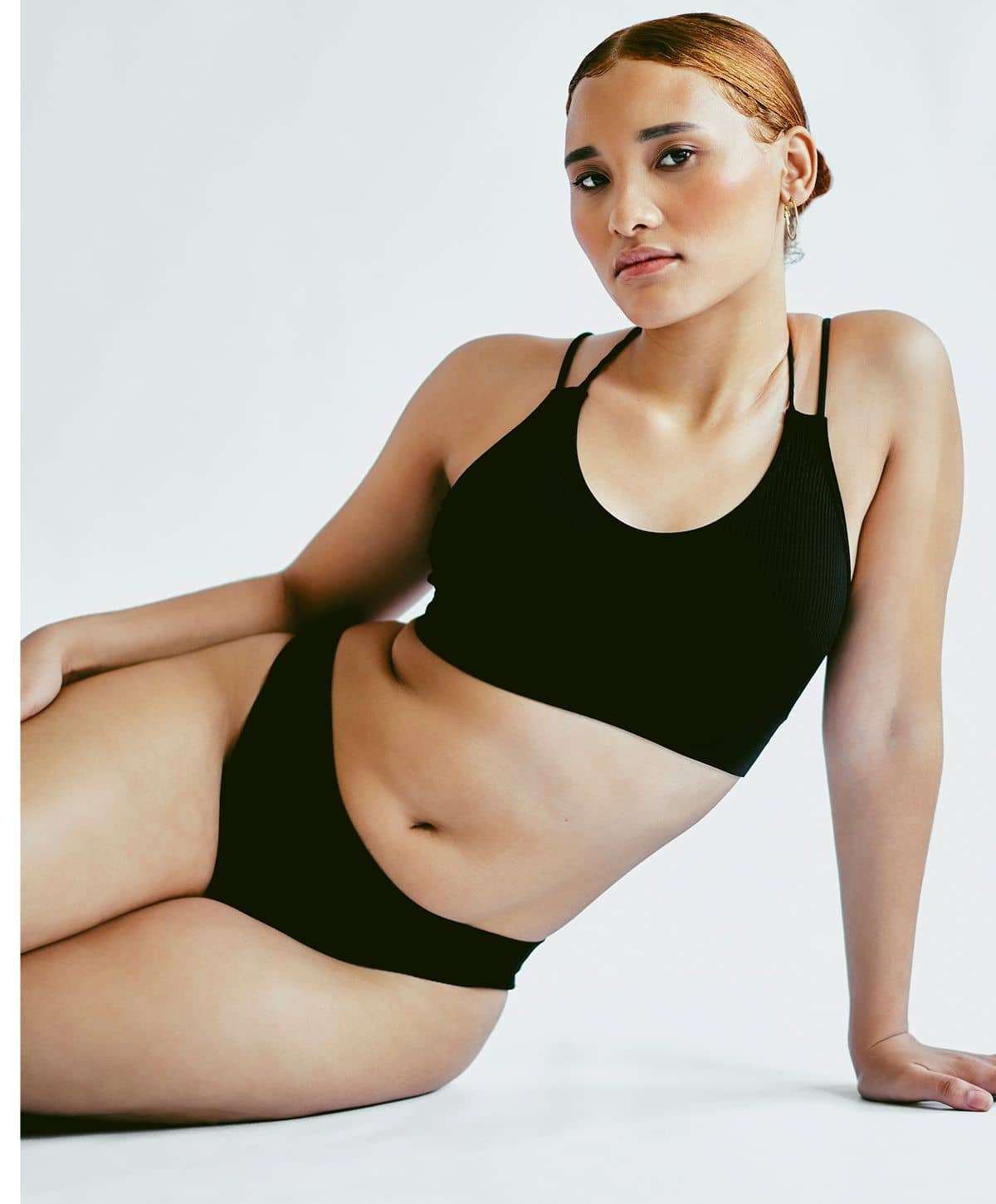 A model reclining in a black sports bra and matching black bikini bottoms, with a subtle smile, showcasing a confident pose.