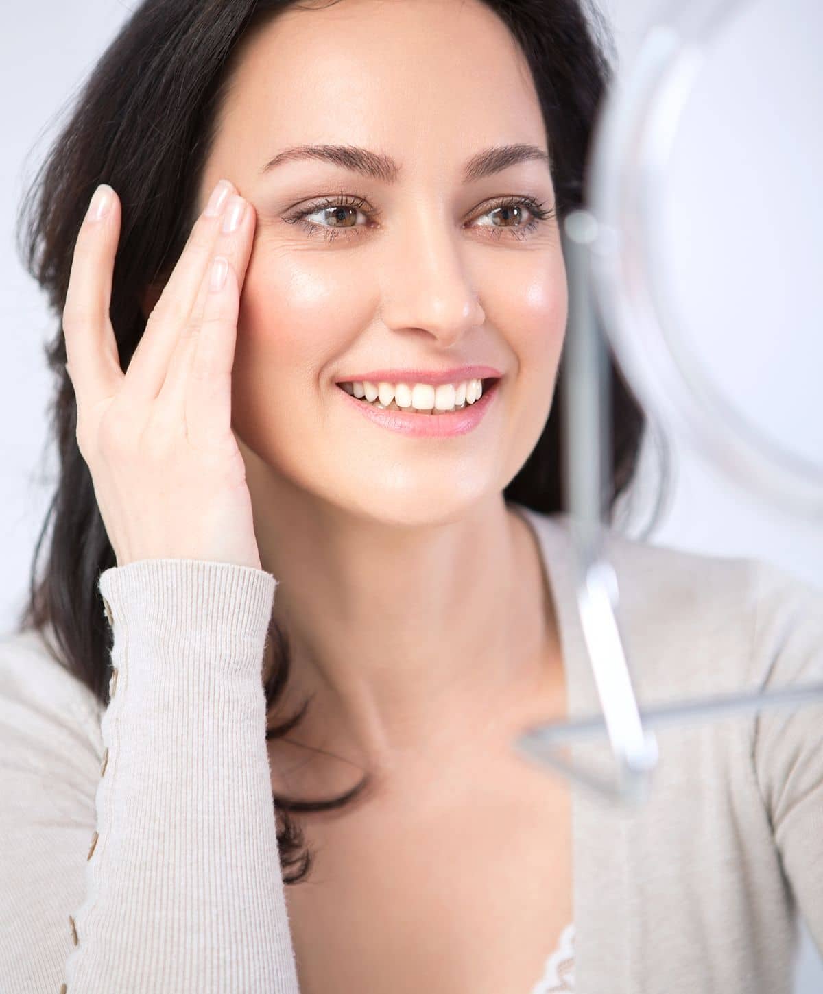 A woman with long dark hair smiling while touching her cheek, looking at her reflection in a handheld mirror.