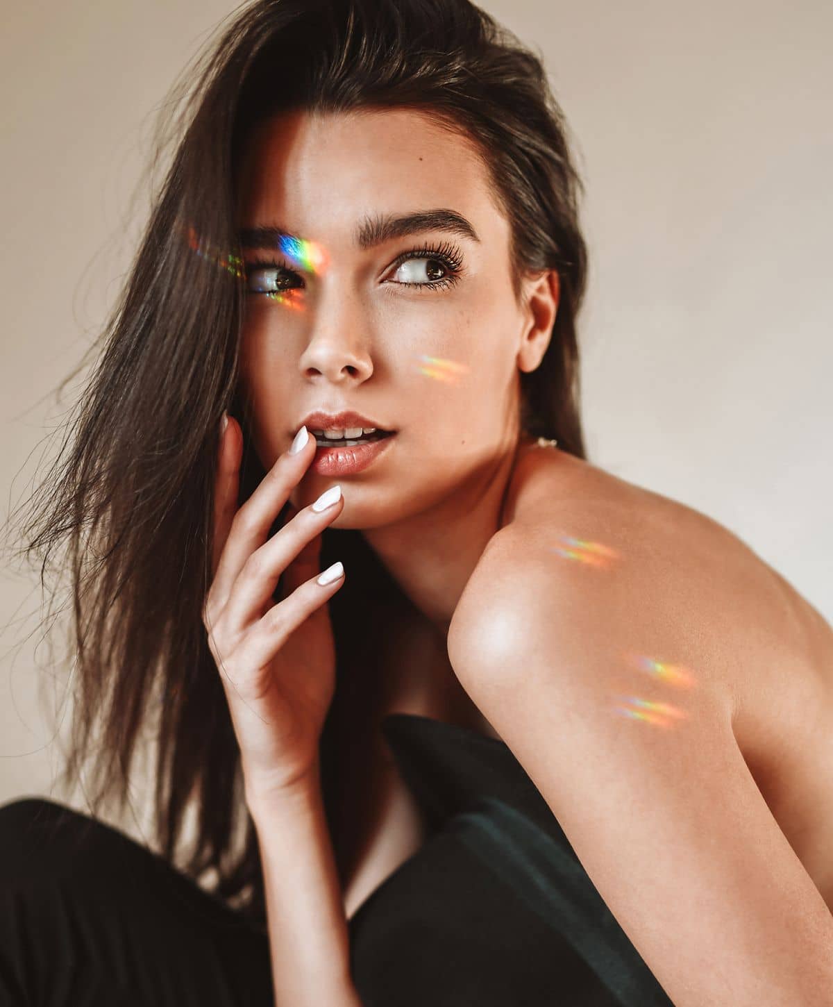 A woman with long dark hair and a natural makeup look, posing with a hand on her cheek, while looking away from the camera. Soft lighting creates subtle highlights on her skin.
