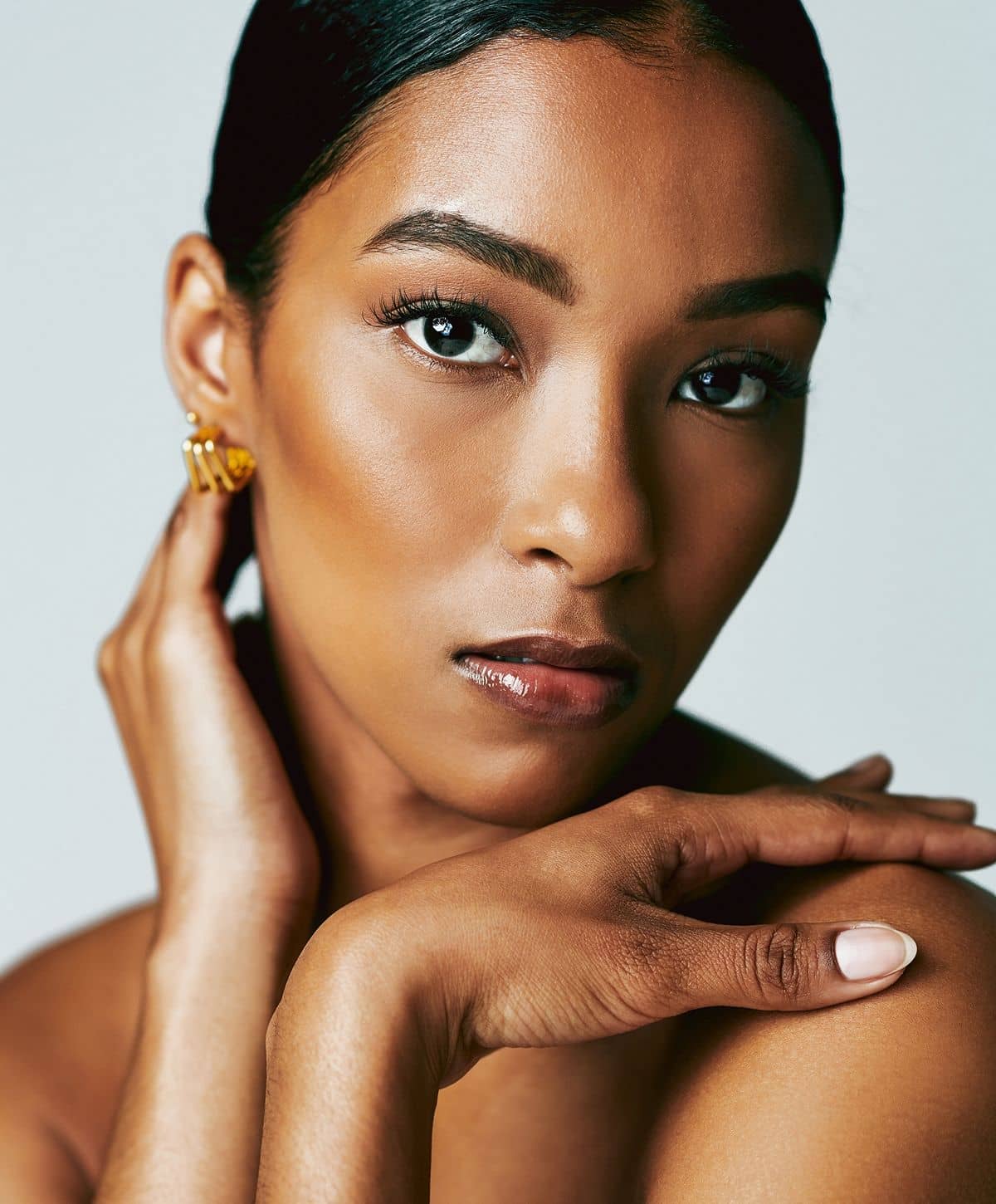 A close-up portrait of a woman with long black hair, wearing gold earrings, gently resting her hand on her neck and shoulder. She has a natural makeup look, highlighting her smooth skin and expressive eyes.