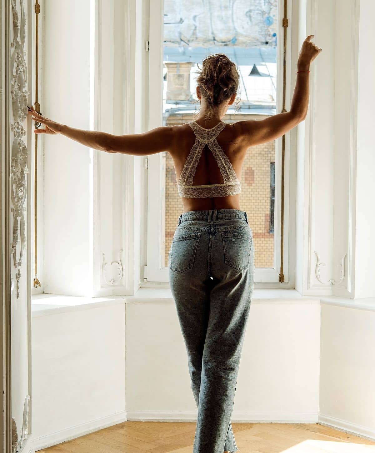 A woman with her back to the camera standing in a window, wearing a lacy, halter-style top and high-waisted jeans, with her arms raised in a relaxed pose.