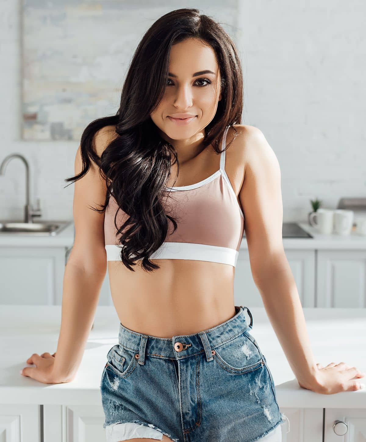 A woman with long dark hair wearing a light pink sports bra and denim shorts, leaning forward on a kitchen counter, smiling at the camera.