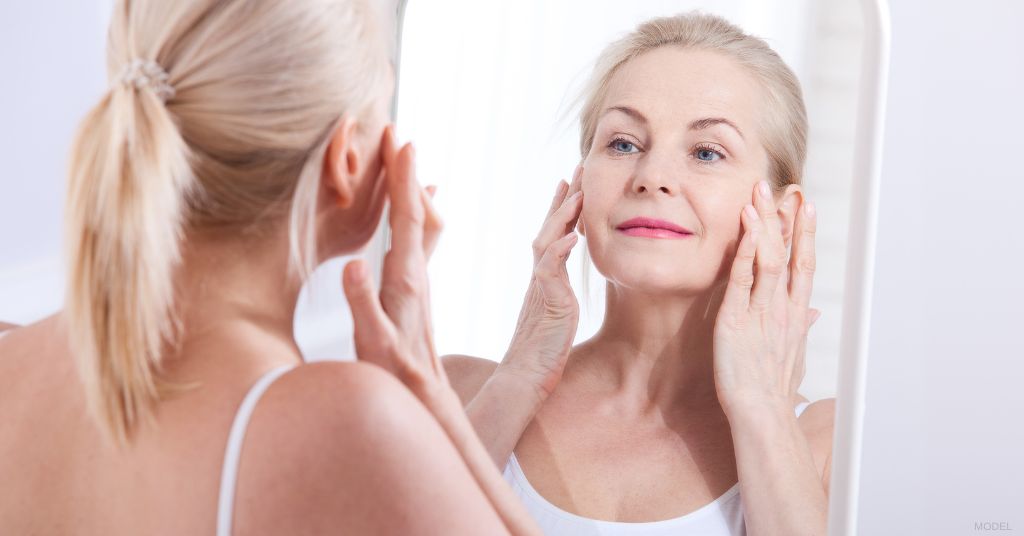 Women after facelift procedure smiling into the mirror and touching her face (model)