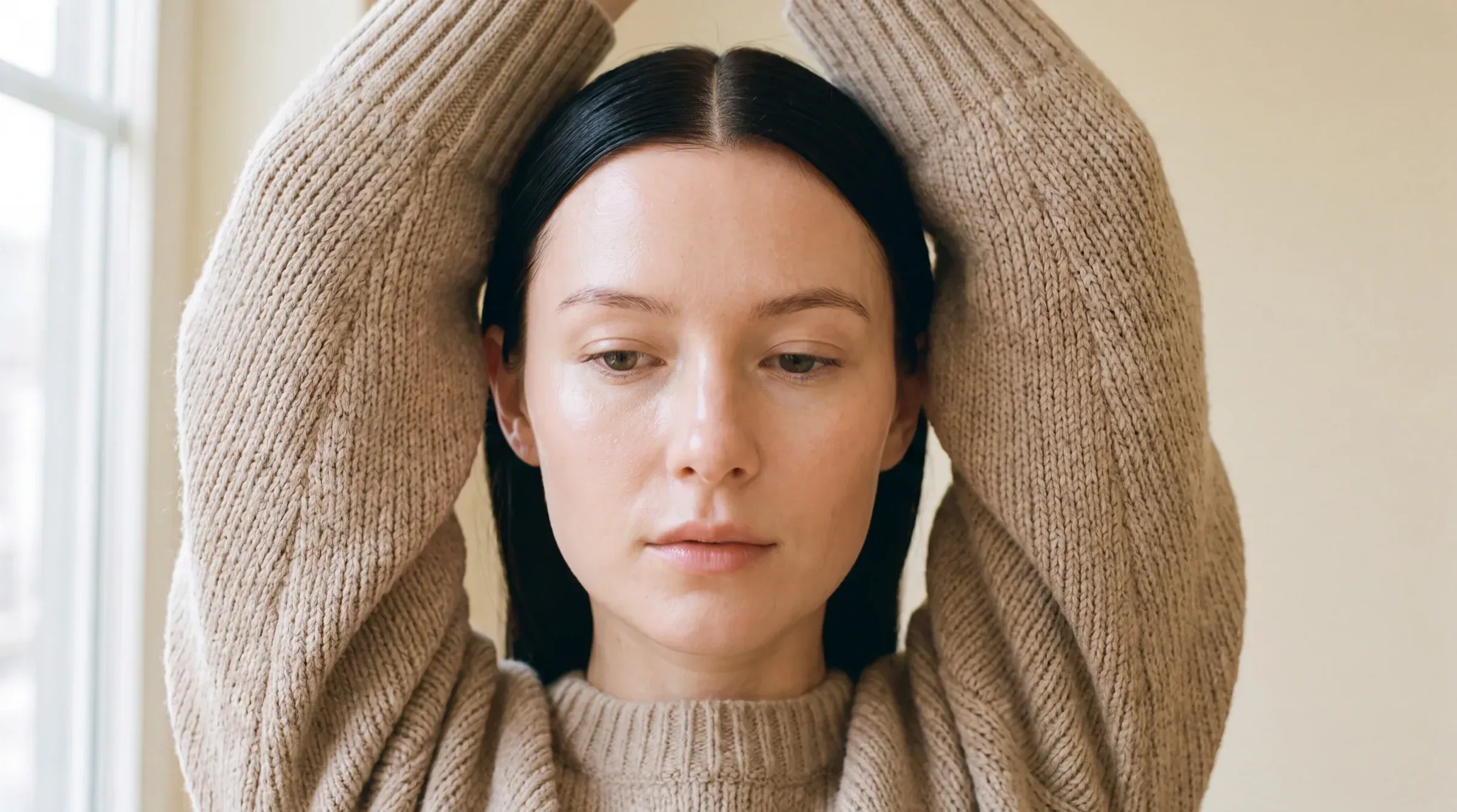 Woman in a beige knit sweater with her arms raised behind her head, in natural light.