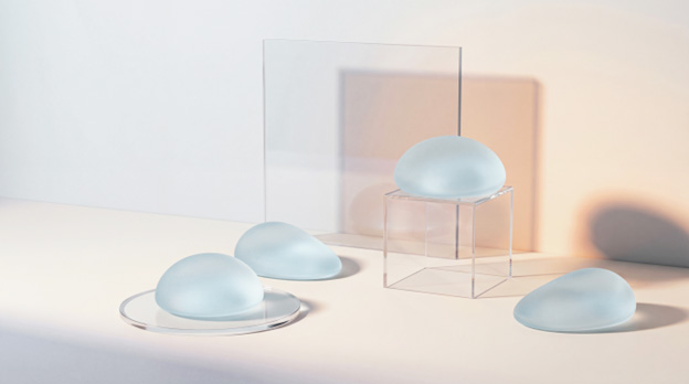 Four frosted silicone breast implant shapes arranged on a clear acrylic display, with varying positions and orientations.