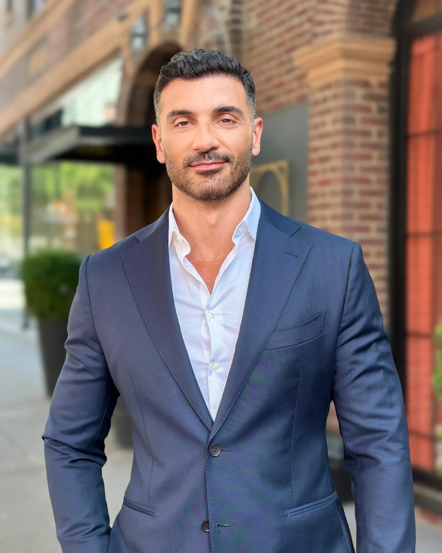 Dr. Antonios Mitsakos in a navy suit and open-collar white shirt, photographed on a city street.