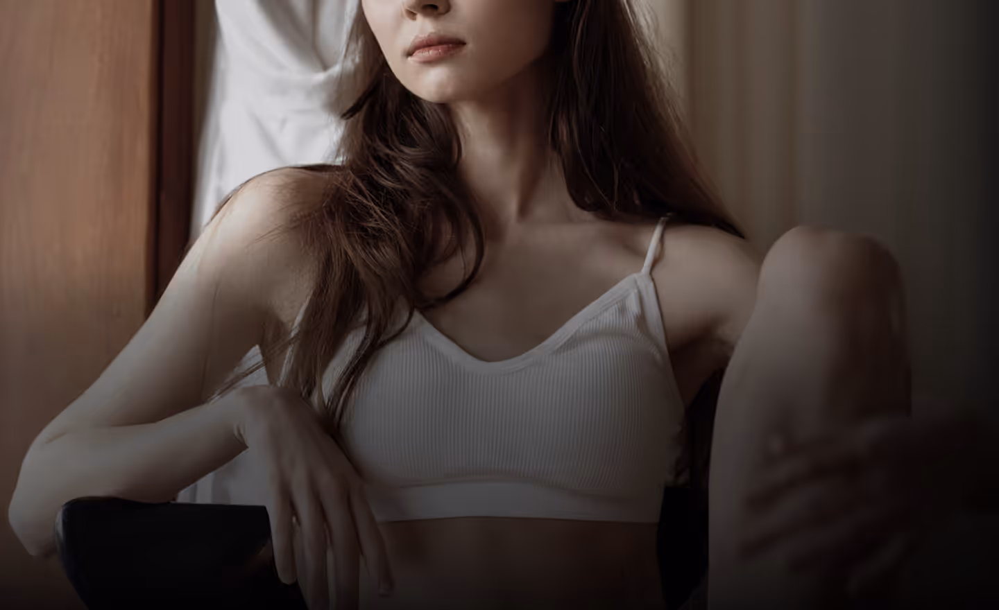 A woman seated, wearing a white bralette, with her hand resting on her hip and her legs crossed.