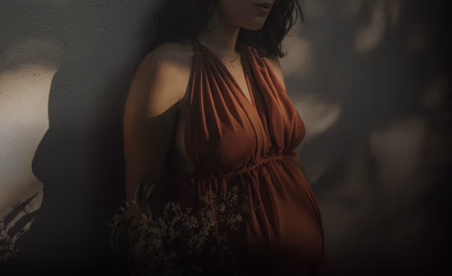 A woman in a rust-colored dress holding a bouquet of white flowers, with a focused expression while standing against a softly lit surface.