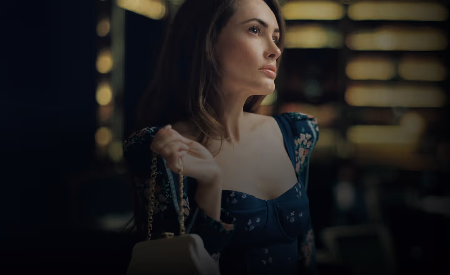 A woman with long brown hair wearing a blue floral dress, holding a white handbag, gazing thoughtfully to her right.