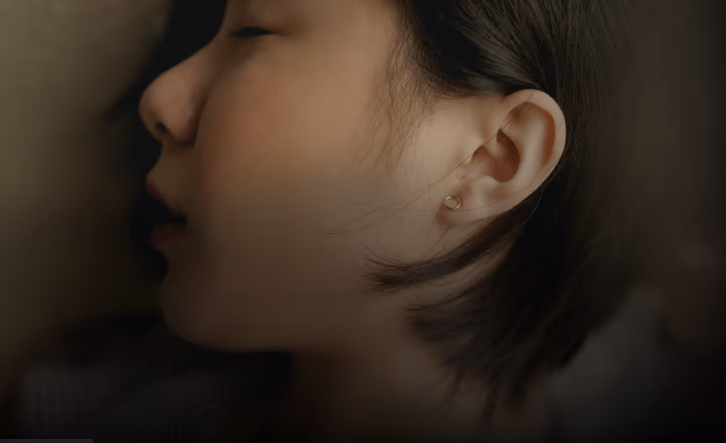 A close-up profile of a young woman with short hair, resting her head against a surface, displaying a small pearl earring in her ear.