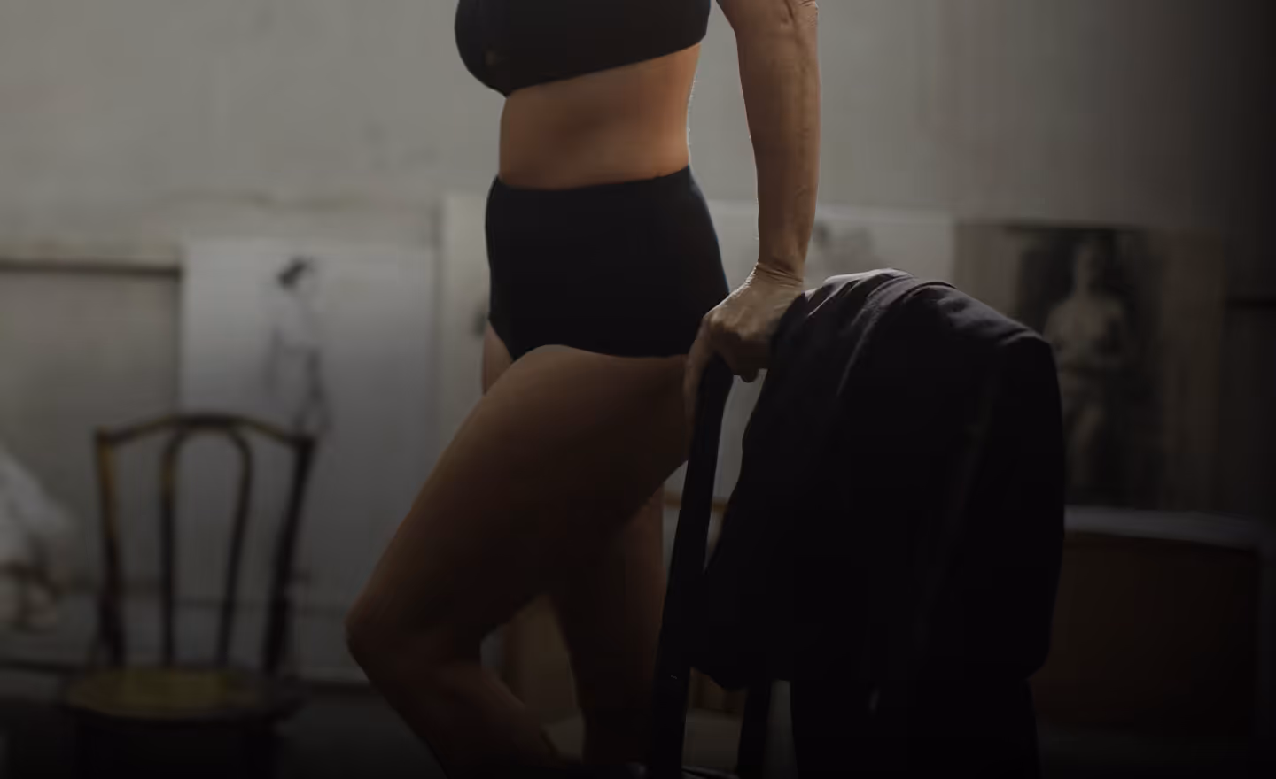 A woman in black high-waisted shorts and a matching cropped top, standing with one hand on a chair and the other holding a black jacket.