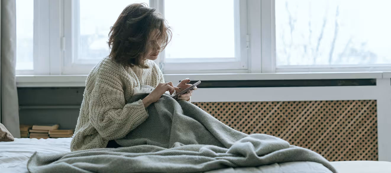 Kvinna i stickad tröja som sitter på en säng och tittar på sin mobiltelefon