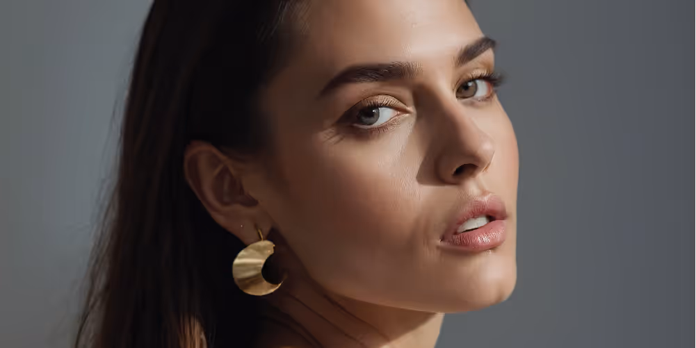 A close-up profile of a woman with long hair and gold circular earrings, showcasing her natural makeup and highlighted features.