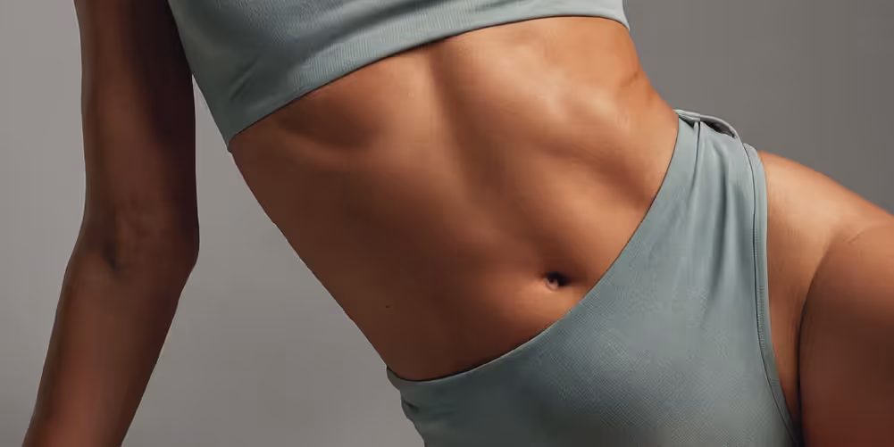 A close-up view of a woman's midsection wearing a greenish-blue crop top and matching high-waisted pants, showcasing toned abs and smooth skin.